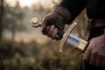 Close up of a longsword hilt with an engraved brass disc pommel and curved brass guard held in a gloved hand, red scabbard at the hip