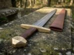 Low angle of a Damascus medieval sword showing the faceted brass pommel and engraved brass cross on a mossy stone bench