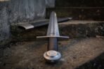 Low angle view of a Damascus bastard sword with wheel pommel and black grip on stone steps beside its scabbard