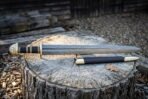 Low side view of a Damascus Viking sword and black scabbard with brass fittings resting on a cut log among wood chips