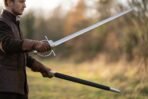 Man holding a steel fencing sword with ring guard and spherical pommel and its black scabbard in a field