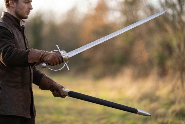 Man holding a steel fencing sword with ring guard and spherical pommel and its black scabbard in a field