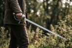 Man in a field jacket holding a cup hilt rapier with a long thin steel blade lowered toward tall grass