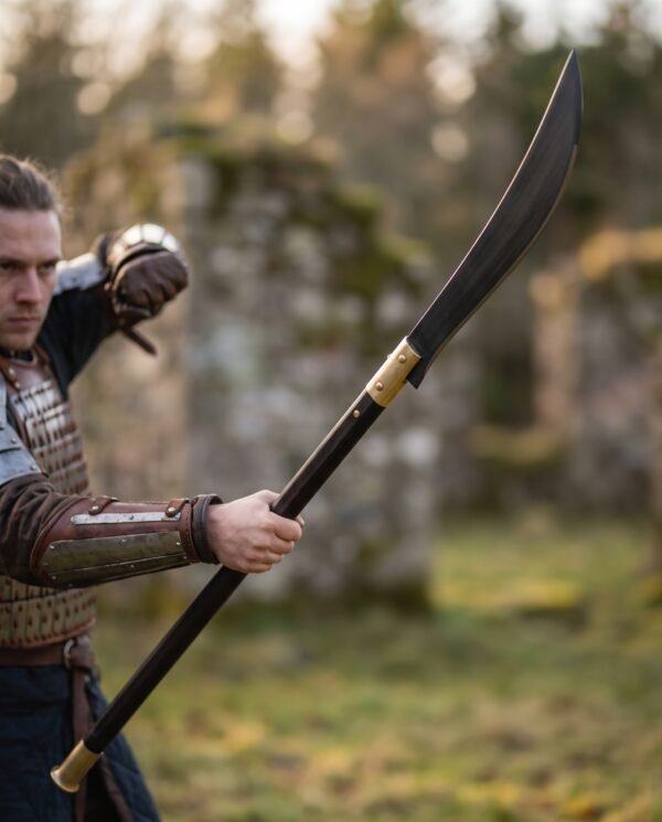 Man in leather armour swinging a medieval glaive with blackened single edged blade and long dark haft in a ruin