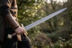 Man in a leather bracer holding a Damascus medieval sword with brass cross and pommel point forward in a sunlit garden
