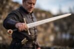 Man in a leather jerkin holding a steel longsword with an engraved inscription and brass hilt out toward the camera