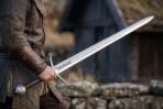 Man in mail and leather holding a polished arming sword by its brown grip in front of a turf roofed timber house