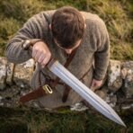 Man in a wool tunic looks down at a Damascus steel gladius held across his body by a dry stone wall, scabbard on his belt