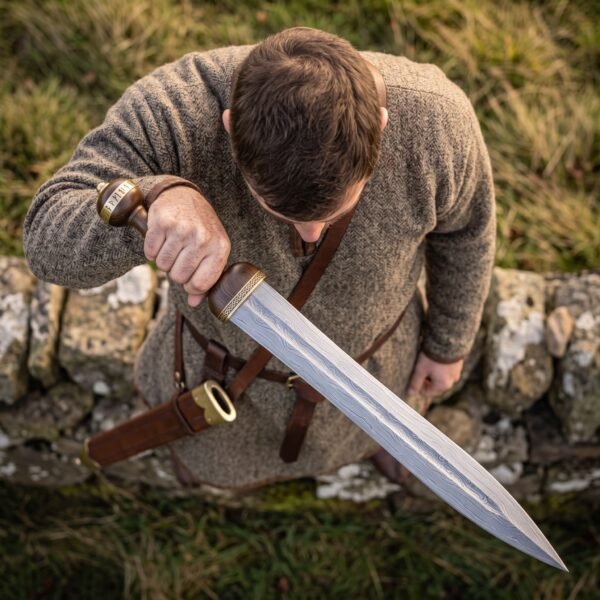Man in a wool tunic looks down at a Damascus steel gladius held across his body by a dry stone wall, scabbard on his belt