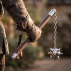 Medieval flail with a walnut haft, polished steel ferrules and a spiked steel ball on a chain, held in a gloved hand outdoors