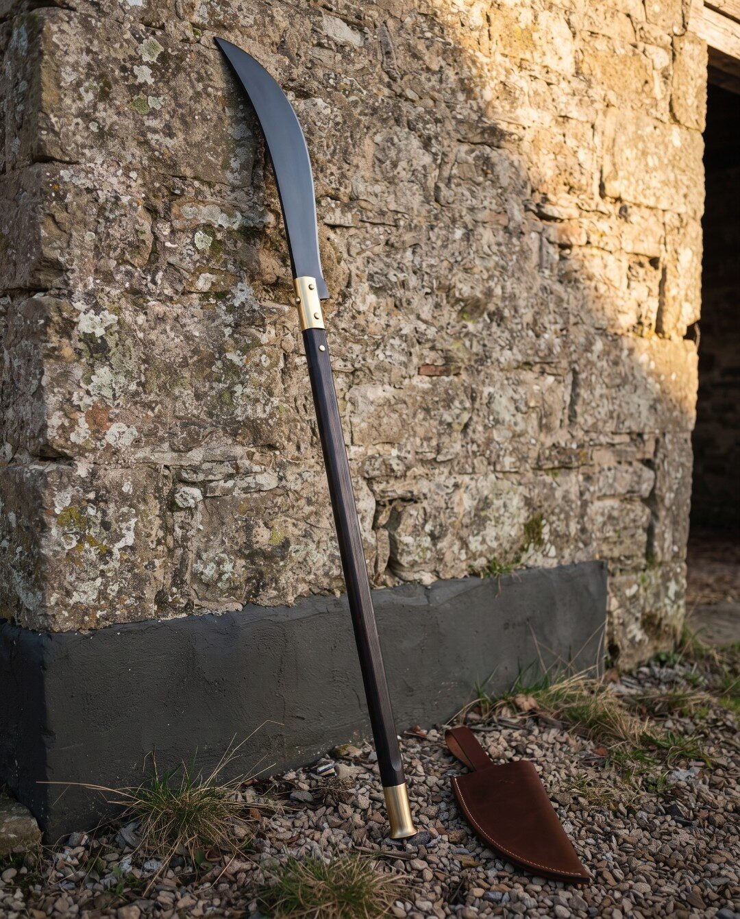 Medieval glaive with a long blackened curved steel blade and brass langets on a dark haft leaning against a rough limest Medieval glaive with a long blackened curved steel blade and brass langets on a dark haft leaning against a rough limestone barn wall on gravel