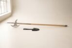 Medieval halberd laid flat on a pale barn floor, satin steel spike and crescent blade on a long ash haft with steel langets beside a black leather head cover