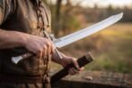 Messer sword held by its dark wooden grip in one hand with the brown leather scabbard in the other, clipped blade tip pointing up