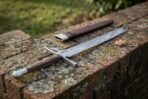 Messer sword laid along a lichen covered brick wall, dark wooden grip with steel pommel cap, brown leather scabbard with steel chape above