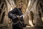 Musketeer rapier with a brass shell guard and swept knuckle bow held point up by a man in a dark doublet under stone vaulted arches