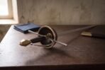 Musketeer rapier with a pierced brass cup hilt and long slender blade laid on a wooden table beside a black scabbard with brass fittings