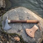 Native American style tomahawk with Damascus head, brass collar and dark walnut haft with leather lacing resting on a river smoothed rock beside its natural leather head cover