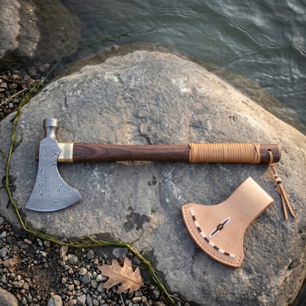 Native American style tomahawk with Damascus head, brass collar and dark walnut haft with leather lacing resting on a river smoothed rock beside its natural leather head cover