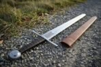 Plain steel arming sword with knotwork etched blade, black leather grip and wheel pommel on gravel beside a tan scabbard
