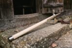 Poleaxe with a pale ash haft and steel butt cap leaning on a stone step by an old timber door, the axe head at the top