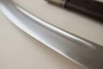 Polished high carbon steel talwar blade, close up of the curve and fuller with the tooled scabbard blurred behind