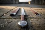 Polished steel arming sword seen from the pommel end, engraved disc pommel and leather grip with scabbard and belt on flagstones