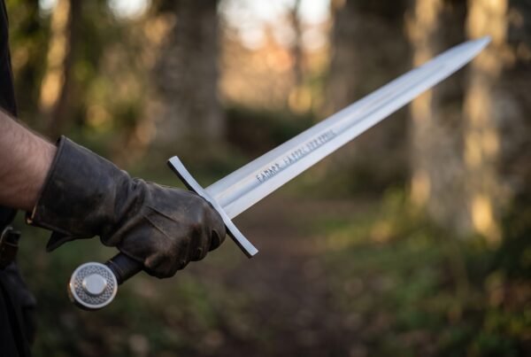 Polished steel arming sword with etched rune line along the fuller held out in a leather gloved hand on a forest path
