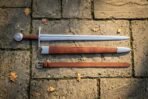 Polished steel arming sword with knotwork disc pommel and russet leather grip laid beside its tan scabbard and belt on stone paving