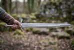 Polished steel elven longsword held level in one hand showing the rune engraved fuller and curved guard in the woods