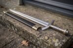 Polished steel elven longsword with engraved rune fuller and ribbed metal grip resting on a stone ledge by a black scabbard