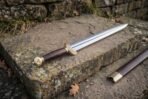 Polished steel fullered sword with wood grip and brass guard lying on a stone step beside its wooden scabbard with brass throat