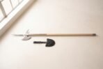 Polished steel halberd with crescent blade and spike on a long pale ash haft lying on a white floor beside a black leather head cover
