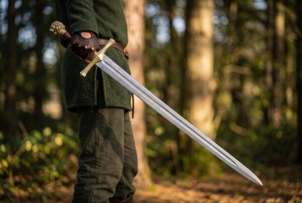 Polished steel longsword with gilt brass cross guard and lion head pommel held at the side of a man in a forest