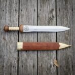 Polished steel Roman gladius shown from above beside its brown leather scabbard with brass throat and chape on wood