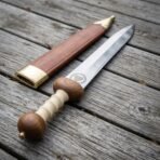 Polished steel Roman gladius with bone grip and wooden pommel lying beside its brown leather scabbard on grey decking