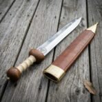 Polished steel Roman gladius with Valknut roundel and ribbed bone grip next to a brass tipped leather scabbard on wood