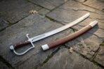 Polished steel sabre with knuckle bow guard and riveted wood grip lying beside its brown leather scabbard on slate paving
