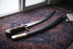 Polished steel shamshir with wood grip and drooping quillons lying with its dark leather scabbard on a red patterned rug