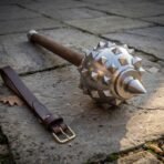 Polished steel spiked mace head close up with pyramid spikes and wood haft lying next to a leather wrist strap on flagstones