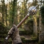 Polished steel spiked mace with conical finial and wood haft gripped in a gloved hand among ruins in the woods