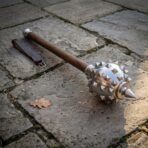 Polished steel spiked mace with steel collars and wood haft resting on worn paving beside a brown leather strap with brass buckle