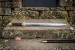 Polished steel straight sword with brass fittings and dark wood grip resting on a stone ledge in front of a dry stone wall