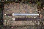 Polished steel straight sword with brass knotwork guard, dark wood grip and round brass pommel above its wooden scabbard on stone