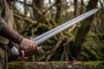 Polished steel Viking broadsword with rune engraved guard and knotwork pommel drawn from a brown leather scabbard in a mossy forest