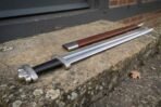 Polished steel Viking sword with black grip and lobed pommel lying on a stone step beside its brown leather scabbard