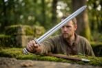 Polished steel Viking sword with a wide fuller held out at arm length by a bearded man in a mossy forest ruin