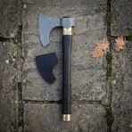 Ragnar axe style hand forged damascus bearded head with a dark hardwood haft and brass collar, resting on flagstone beside its black leather head sheath