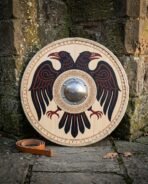 Ravenward hand made Viking round shield with hand painted black and red raven motif, Elder Futhark rune border and hand forged steel boss leaning against a stone wall