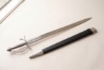 Renaissance short sword with a straight fullered blade and side ring guard laid on a pale surface with its black leather scabbard
