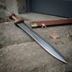 Roman gladius with ribbed wood grip and round pommel lying on a stone step beside its tooled leather scabbard with brass throat